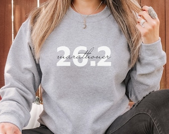 May include: A gray sweatshirt with the text "26.2 marathoner" printed in white on the front. The sweatshirt is being worn by a person sitting on a checkered blanket with pumpkins in the background.