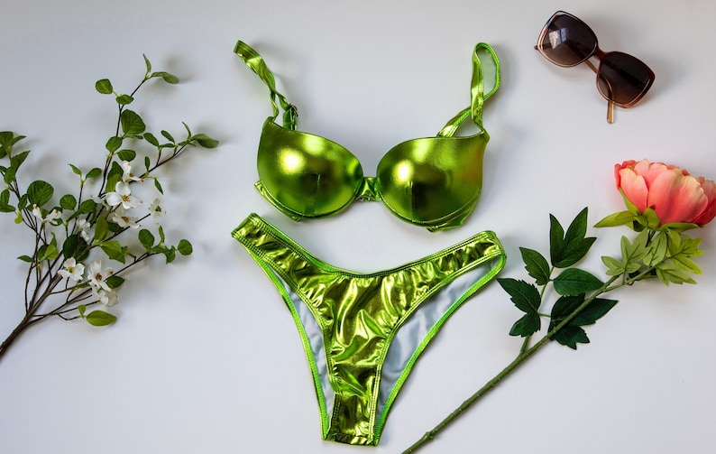 May include: A green metallic bikini with a push-up bra style top and a high-cut bottom. The bikini is on a white background with a flower and a branch of white flowers.