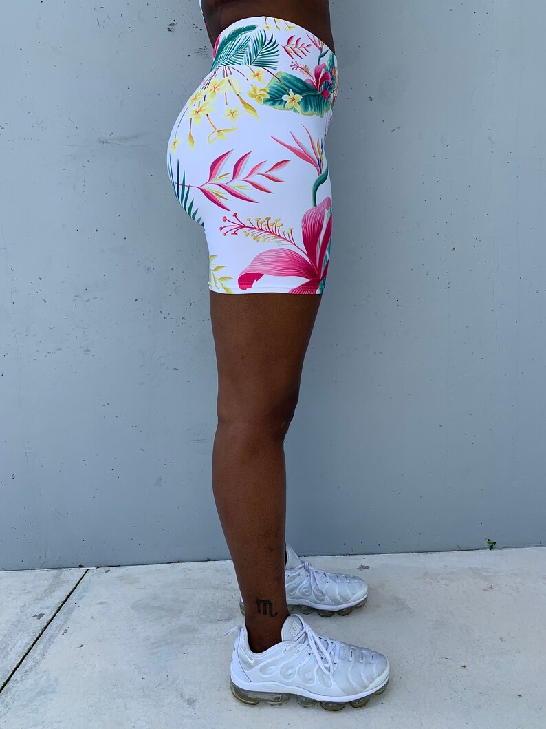 May include: White biker shorts with a vibrant floral pattern featuring pink, red, yellow, and green tropical flowers and leaves. The shorts are form-fitting and high-waisted, perfect for workouts or casual wear.