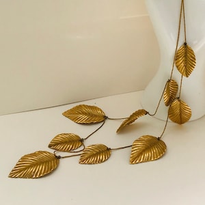 May include: A gold necklace with multiple strands of chain and leaf-shaped pendants. The necklace is displayed on a white mannequin hand.