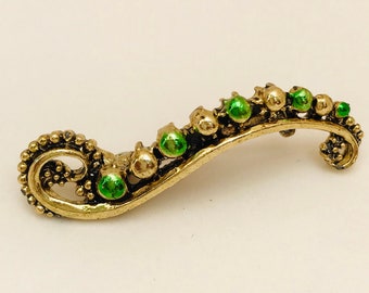 May include: A gold-colored brooch with a curved design and green enamel accents. The brooch is shaped like a stylized leaf or branch with a series of small green circles.