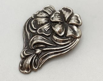 May include: A silver-toned brooch in the shape of a stylized flower with intricate details. The flower has five petals and is surrounded by swirling leaves.