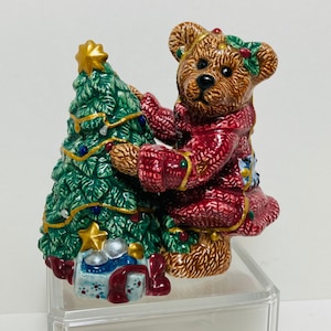 May include: A ceramic figurine of a brown teddy bear wearing a red sweater and holding a green Christmas tree with gold ornaments. The figurine is sitting on a clear acrylic base.