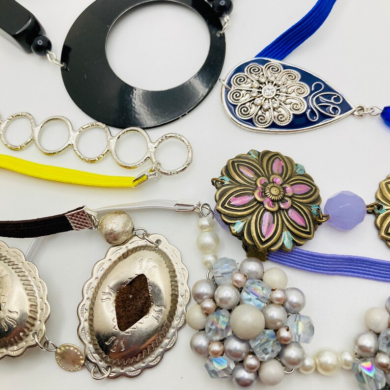 May include: A collection of colorful and silver jewelry pieces, including a black circle pendant, a blue and silver teardrop pendant, a silver flower pendant, a silver and brown pendant with a floral design, and a cluster of white and iridescent beads.