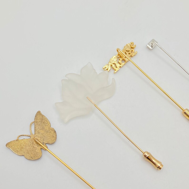 May include: An assortment of decorative pins with various designs. The pins include a gold butterfly, a frosted white flower, a gold lion, and a silver square-topped pin. The pins are set against a white background.
