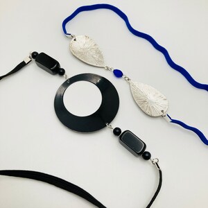 May include: A black and silver necklace with a large black circle pendant, two silver leaf-shaped pendants, and black and blue beads. The necklace is on a white background.
