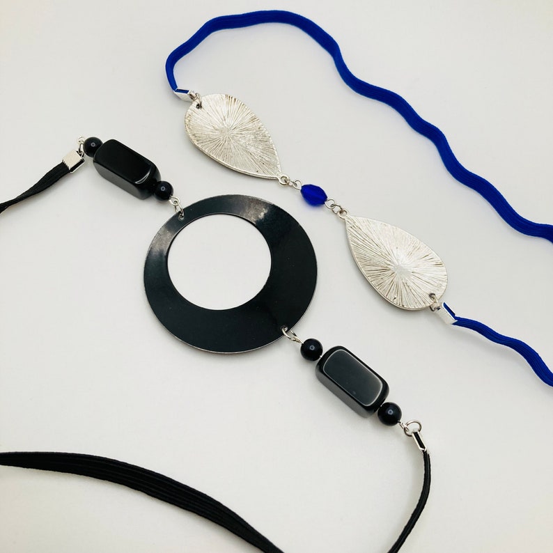 May include: A black and silver necklace with a large black circle pendant, two silver leaf-shaped pendants, and black and blue beads. The necklace is on a white background.