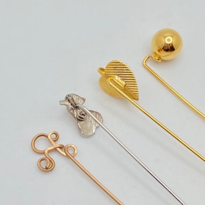 May include: Four decorative hat pins in gold and silver tones. The pins feature various designs, including a ball, a leaf, a swirl, and a small figure. The pins are arranged diagonally against a white background, showcasing their details and craftsmanship.