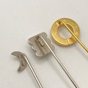 Pode incluir: Close-up de tr&ecirc;s alfinetes decorativos. Um &eacute; um c&iacute;rculo dourado, outro &eacute; uma lua crescente prateada e o terceiro &eacute; um quadrado prateado com uma borda ondulada. Cada alfinete tem um alfinete longo e fino preso na parte de tr&aacute;s.