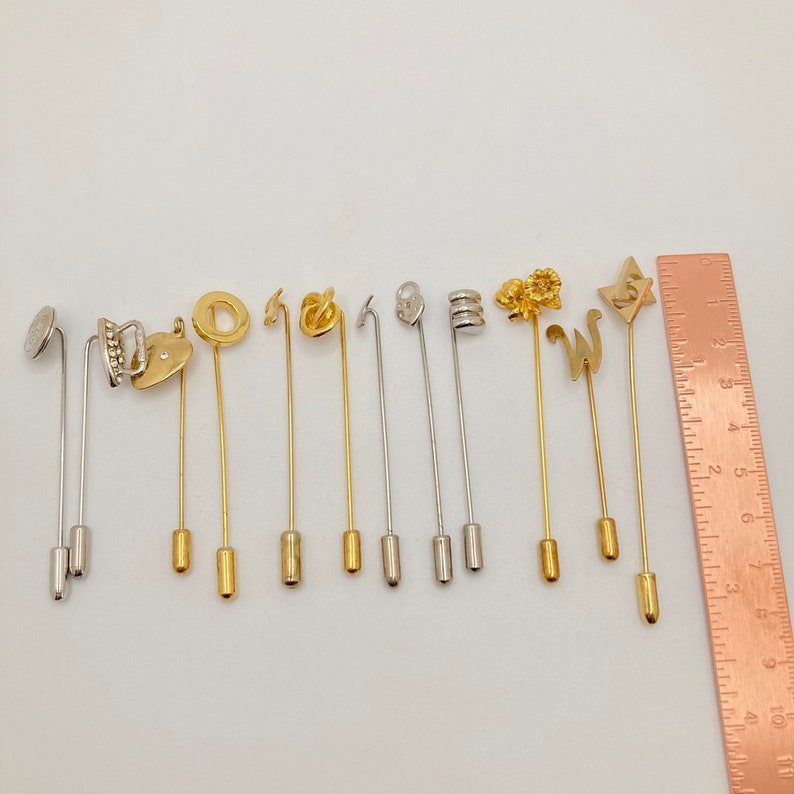Pode incluir: Uma cole&ccedil;&atilde;o de alfinetes de lapela decorativos em tons de ouro e prata. Os alfinetes apresentam v&aacute;rios designs, incluindo um cora&ccedil;&atilde;o, um c&iacute;rculo e a letra "W". Uma r&eacute;gua &eacute; colocada ao lado dos alfinetes para escala.