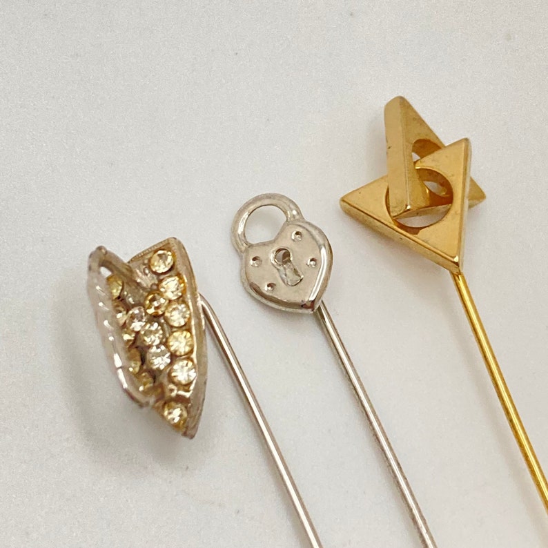 Pode incluir: Tr&ecirc;s alfinetes decorativos para chap&eacute;us. Um alfinete apresenta um design de cadeado em forma de cora&ccedil;&atilde;o prateado. Outro alfinete tem um design de tri&acirc;ngulo dourado. O terceiro alfinete tem um design curvo prateado com pequenas pedras preciosas transparentes.