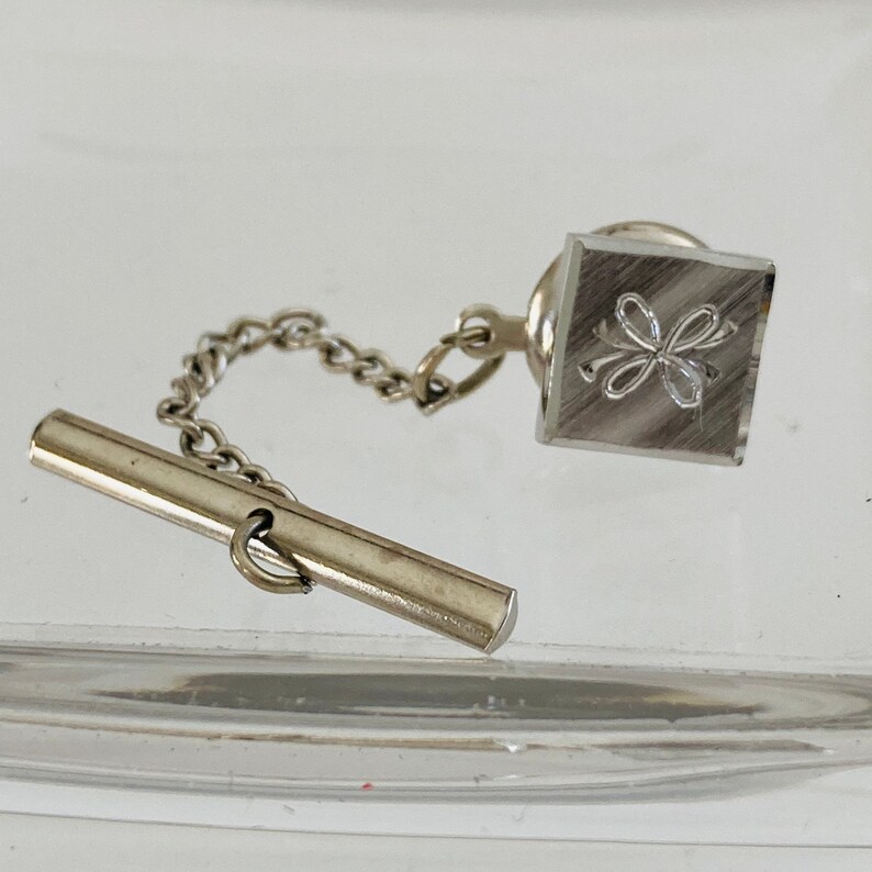 May include: Silver tie tack with a chain and a bar. The tie tack has a square design with a four-leaf clover design.