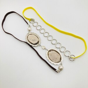 May include: A brown and yellow headband with a silver chain and two oval-shaped silver charms. The charms have a textured surface and a pearl-like bead hangs from the top charm.
