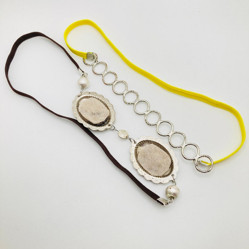 May include: A brown and yellow headband with a silver chain and two oval-shaped silver charms. The charms have a textured surface and a pearl-like bead hangs from the top charm.