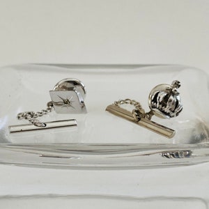 May include: Two silver tie tacks with chains. One features a star with a small diamond in the center. The other features a crown.