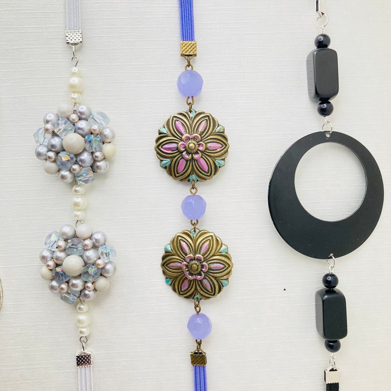 May include: Three decorative hanging ornaments with different styles. The first ornament features a white and silver beaded cluster with a white tassel. The second ornament has a blue and pink floral design with a blue tassel. The third ornament has a black circle with a black square and a black tassel.