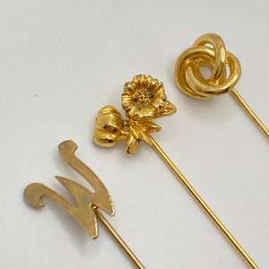 Pode incluir: Tr&ecirc;s alfinetes de chap&eacute;u dourados. Um alfinete apresenta um design floral, outro tem um design de n&oacute; e o terceiro tem um design estilizado da letra "W". Os alfinetes t&ecirc;m hastes longas e finas.