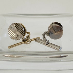 May include: Two silver tie tacks with chain and bar attachments. One tack has a striped design and the other has a geometric design.