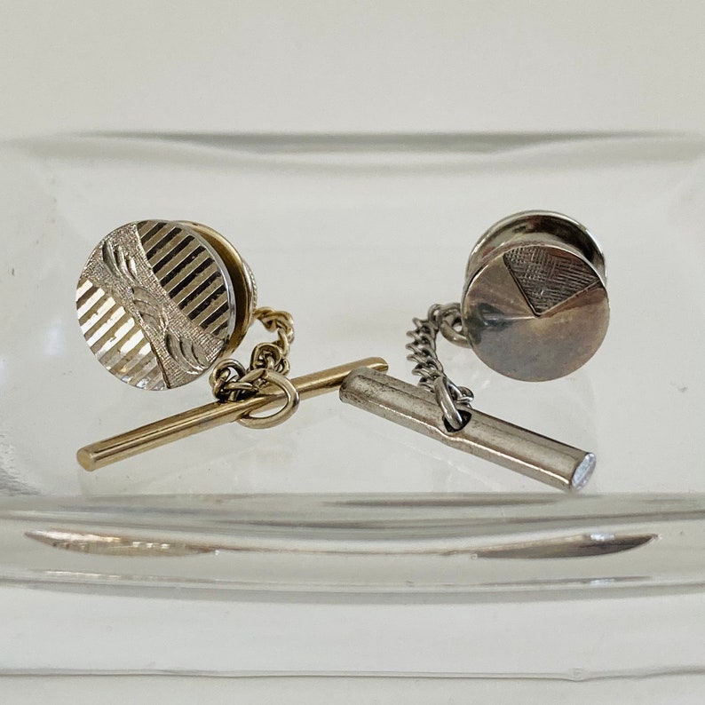 May include: Two silver tie tacks with chain and bar attachments. One tack has a striped design and the other has a geometric design.