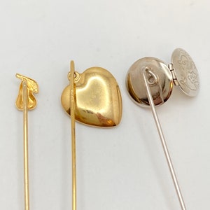 Pode incluir: Tr&ecirc;s alfinetes de chap&eacute;u vintage. Um alfinete dourado apresenta um design em forma de p&ecirc;ra. Outro alfinete dourado tem um pingente em forma de cora&ccedil;&atilde;o. O terceiro alfinete &eacute; prateado com um pingente redondo que se abre para revelar um design floral.
