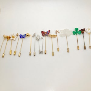 May include: A collection of decorative hat pins with various designs. The pins feature floral, butterfly, and abstract shapes in gold, silver, and colorful enamel. Each pin has a long, slender shaft and a decorative head.