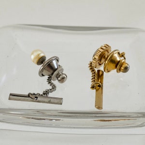May include: Two tie tacks with chains. One is silver with a pearl and the other is gold with a decorative design.