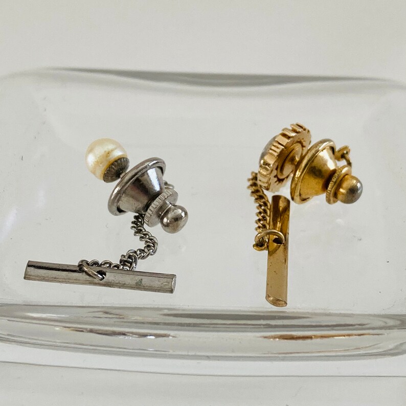 May include: Two tie tacks with chains. One is silver with a pearl and the other is gold with a decorative design.