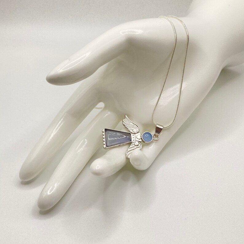 Op de afbeelding: Een zilveren engel hanger ketting met een blauwe edelsteen. De engel heeft gedetailleerde vleugels en een lichaam van helder glas.