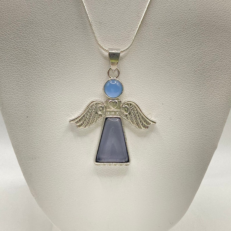 Op de afbeelding: Een zilveren engel hanger ketting met een lichtblauwe en paarse edelsteen. De engel heeft vleugels en een hart op zijn borst.