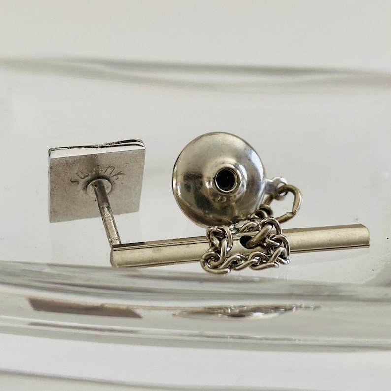 May include: Silver tie tack with a square base and a round top. The base has the word "Sterling" engraved on it. The top has a small black stone in the center.