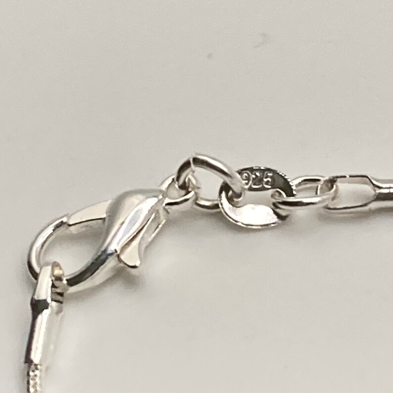 Op de afbeelding: Close-up van een zilveren kettinghalsband sluiting met een karabijn sluiting. De ketting is gemaakt van kleine, ronde schakels. De sluiting is gemarkeerd met 925, wat aangeeft dat het van sterling zilver is.