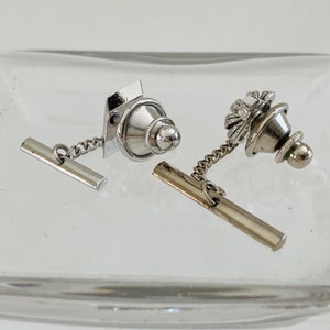 May include: Two silver tie tacks with chains. One tie tack has a round top with a decorative design. The other tie tack has a square top with a round top.