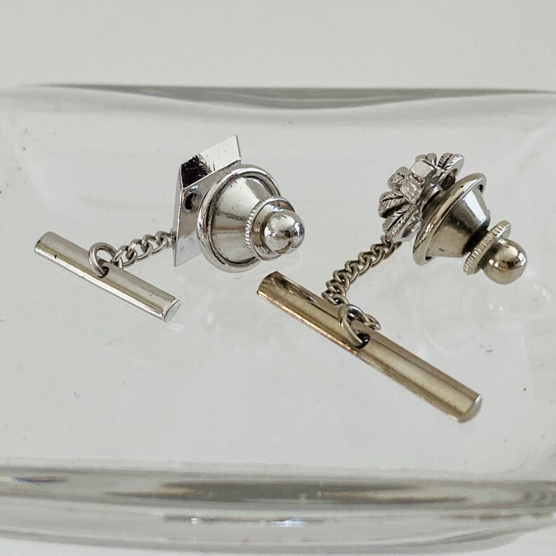 May include: Two silver tie tacks with chains. One tie tack has a round top with a decorative design. The other tie tack has a square top with a round top.