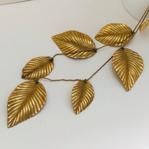 May include: A gold leaf garland with six leaves connected by a thin gold cord. The leaves are textured and have a slightly raised design.
