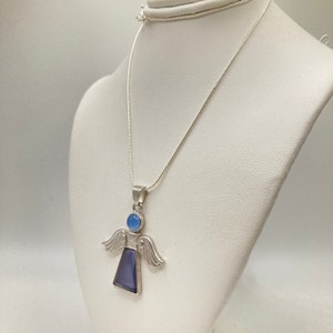Op de afbeelding: Een zilveren ketting met een engelhanger met een blauwe edelsteen. De engel heeft vleugels en een rond hoofd met een blauwe edelsteen.