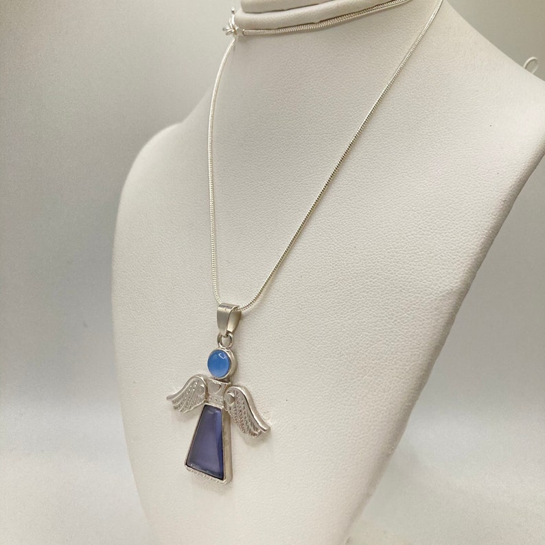 Op de afbeelding: Een zilveren ketting met een engelhanger met een blauwe edelsteen. De engel heeft vleugels en een rond hoofd met een blauwe edelsteen.