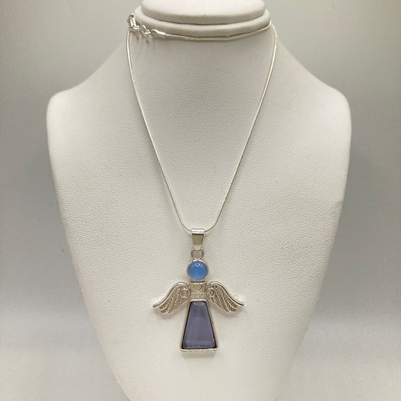 Op de afbeelding: Een zilveren engel hanger ketting met een lichtblauwe en paarse edelsteen. De engel heeft vleugels en een hart op zijn borst.