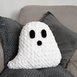 Puede incluir: Almohada fantasma de crochet blanco con ojos de bot&oacute;n negro y boca de bot&oacute;n negro. La almohada est&aacute; sentada en un sof&aacute; gris.