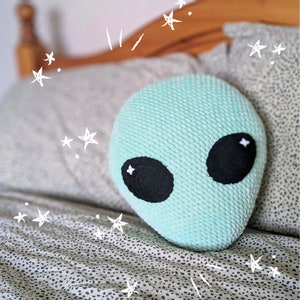 May include: A light teal, crocheted alien head pillow with large black oval eyes and white highlights. The pillow is on a bed with a patterned comforter and decorative stars.
