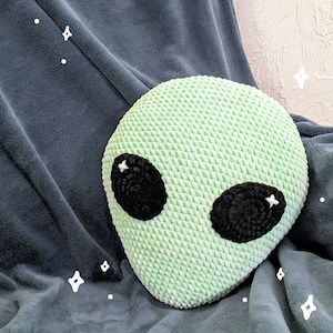 May include: A light green, crocheted alien head pillow with large black eyes and white accents. The pillow is round and sits on a dark gray blanket. The pillow is a fun, decorative item.