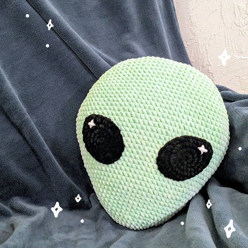 May include: A light green, crocheted alien head pillow with large black eyes and white accents. The pillow is round and sits on a dark gray blanket. The pillow is a fun, decorative item.