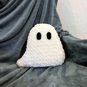 Puede incluir: Una almohada fantasma de crochet blanco con ojos negros. La almohada est&aacute; sobre una manta gris.