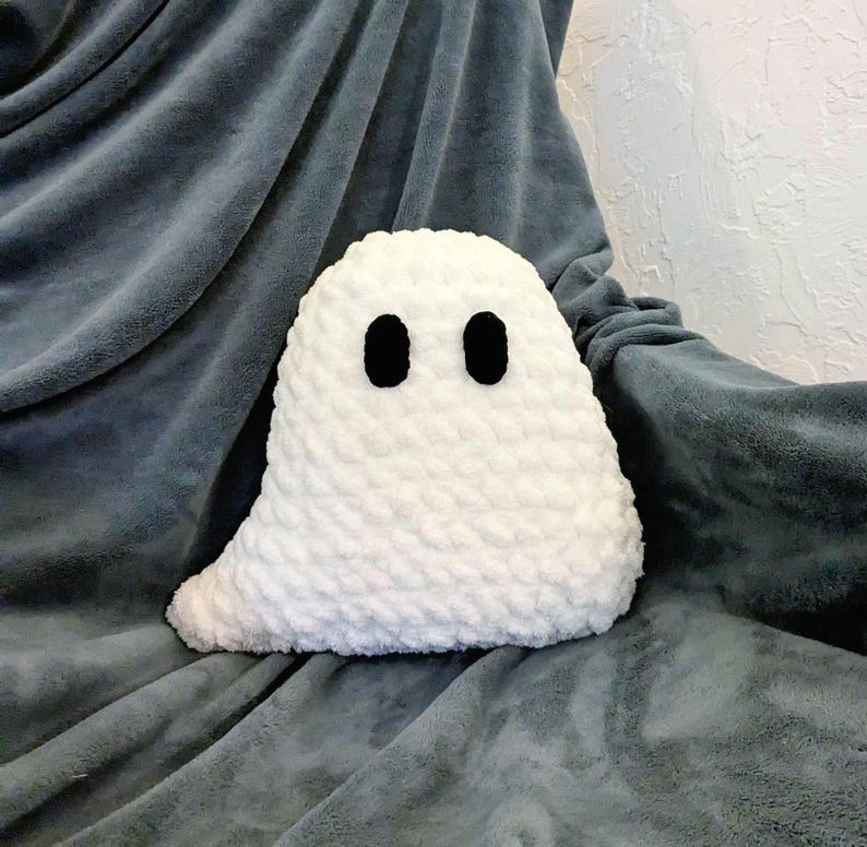 Puede incluir: Una almohada fantasma de crochet blanco con ojos negros. La almohada est&aacute; sobre una manta gris.