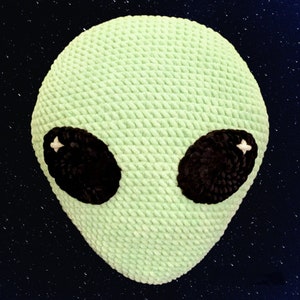 May include: A light green, crocheted alien head with large, oval black eyes. The eyes have white star-shaped highlights. The plush, textured design gives the alien a soft, friendly appearance. The background is a dark blue with white star-like specks.