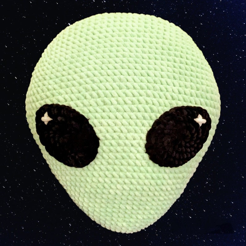 May include: A light green, crocheted alien head with large, oval black eyes. The eyes have white star-shaped highlights. The plush, textured design gives the alien a soft, friendly appearance. The background is a dark blue with white star-like specks.