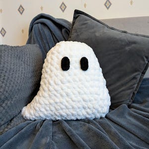 Puede incluir: Una almohada fantasma blanca de crochet con ojos de bot&oacute;n negros, sentada en un sof&aacute; gris.
