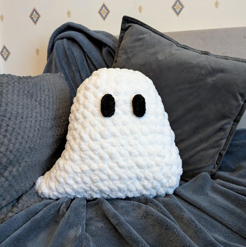 Puede incluir: Una almohada fantasma blanca de crochet con ojos de bot&oacute;n negros, sentada en un sof&aacute; gris.