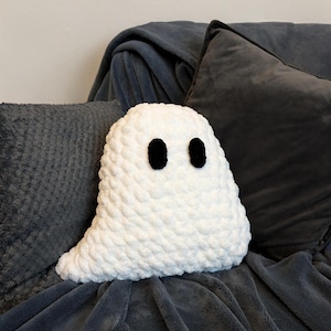 Puede incluir: Una almohada fantasma blanca de crochet con ojos negros, sentada en un sof&aacute; gris.