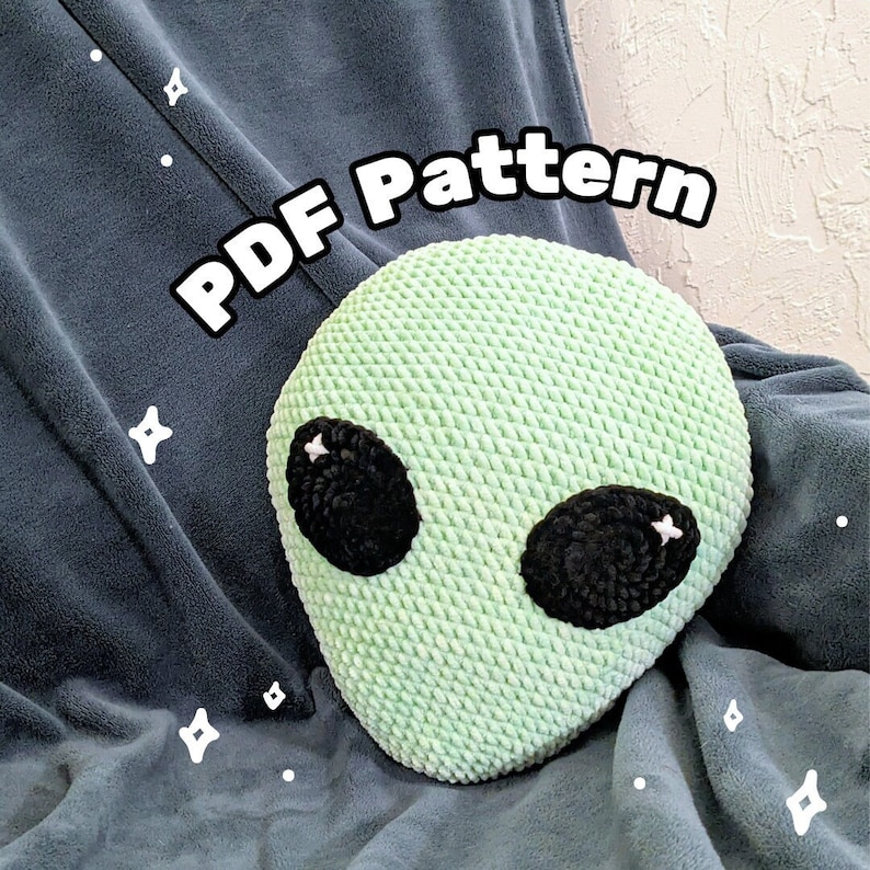 May include: A light green, crocheted alien head with large black eyes and white highlights. The text "PDF Pattern" is displayed in white across the top. The alien is resting on a dark blue blanket.