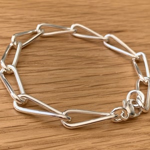 May include: A silver bracelet with a unique chain link design. The links are elongated and teardrop-shaped, connected by smaller, rounded links. The bracelet is secured with a lobster clasp. The jewelry is displayed on a wooden surface.